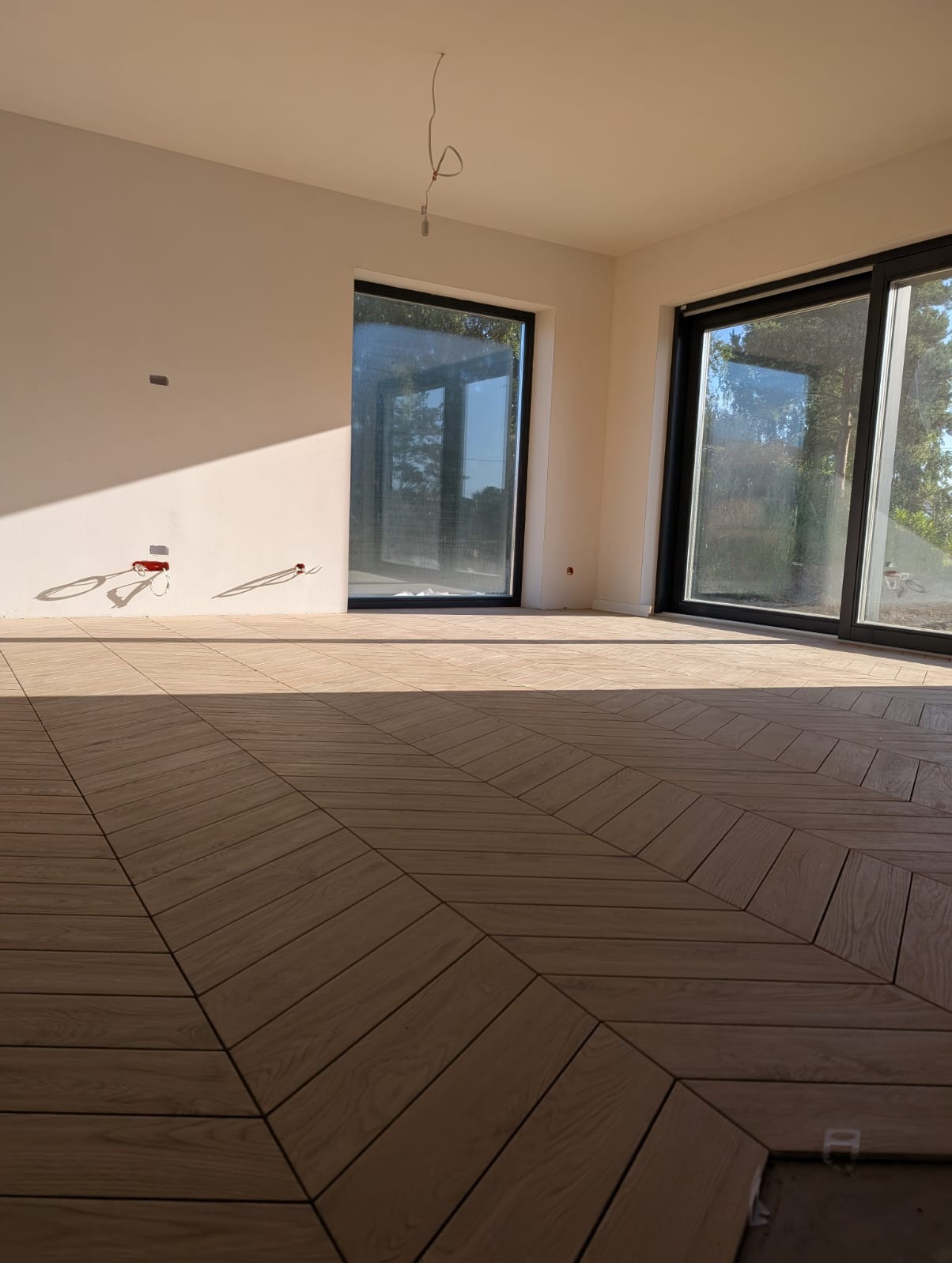 Jasne płytki podłogowe ułożone we wzór jodełki w przestronnym, słonecznym wnętrzu z dużymi oknami. Widoczne niedokończone prace przy krawędzi podłogi.