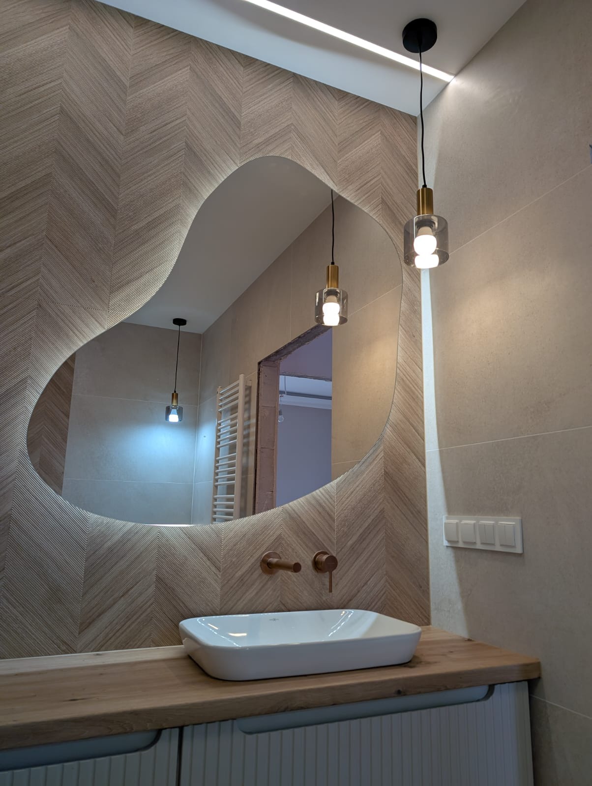 Nowoczesna łazienka z asymetrycznym lustrem podświetlanym LED, drewnianym blatem, białą umywalką i armaturą w kolorze miedzi. Na ścianie płytki ułożone w jodełkę.