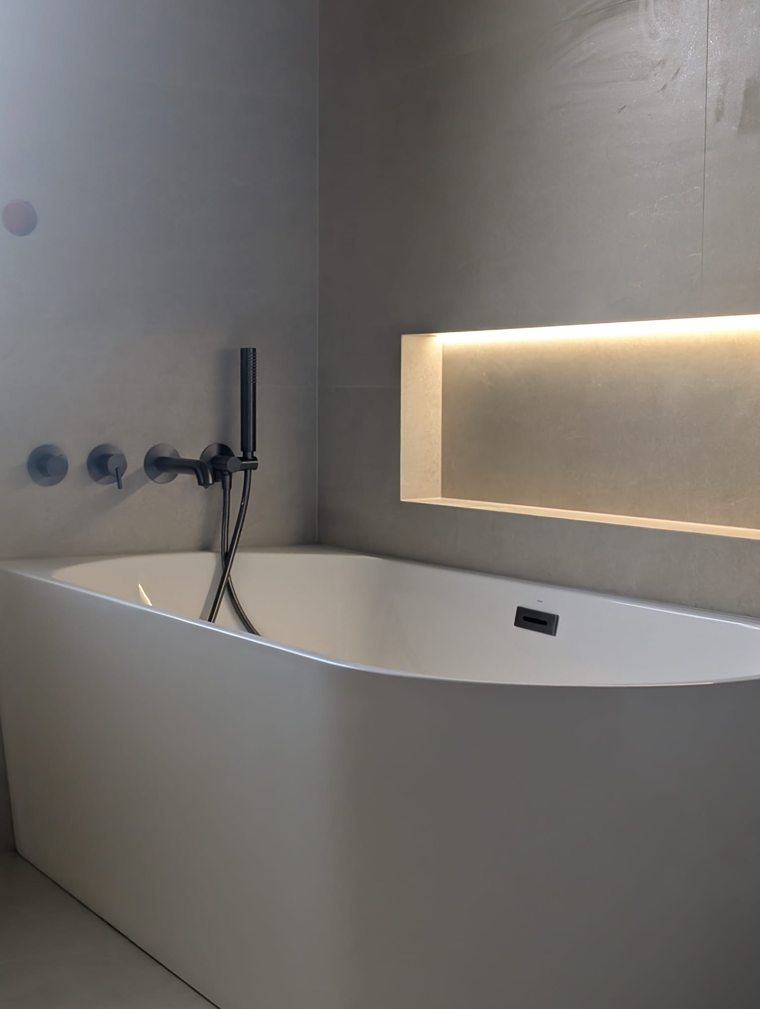 Minimalistyczna łazienka: biała wanna wolnostojąca z czarną armaturą na tle szarej ściany z podświetloną wnęką. Nowoczesny design, stonowana kolorystyka.