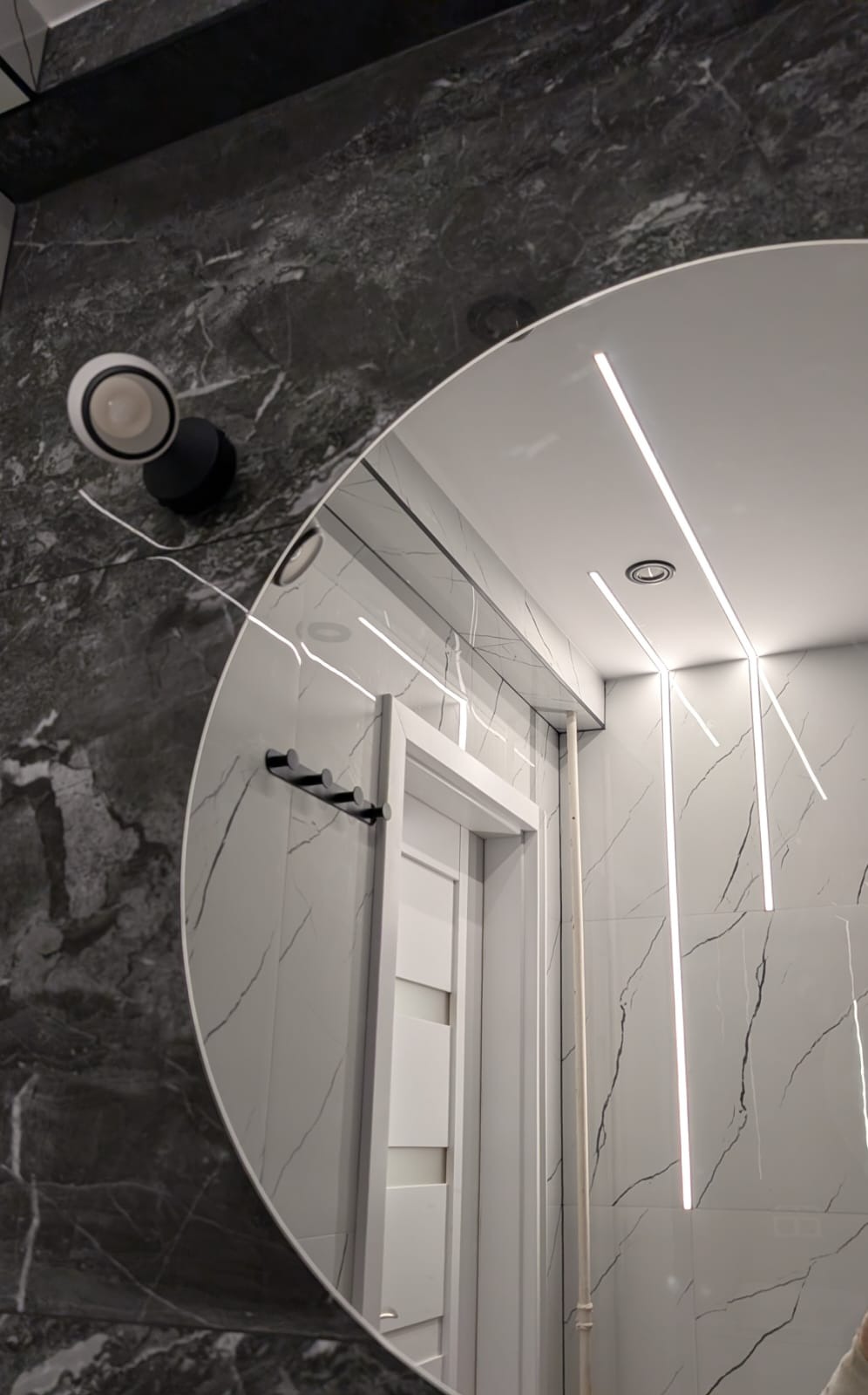 Okrągłe lustro odbija nowoczesną łazienkę z marmurowymi płytkami, oświetleniem LED i białymi drzwiami. Czarny wieszak na ręczniki i lampa dodają kontrastu.