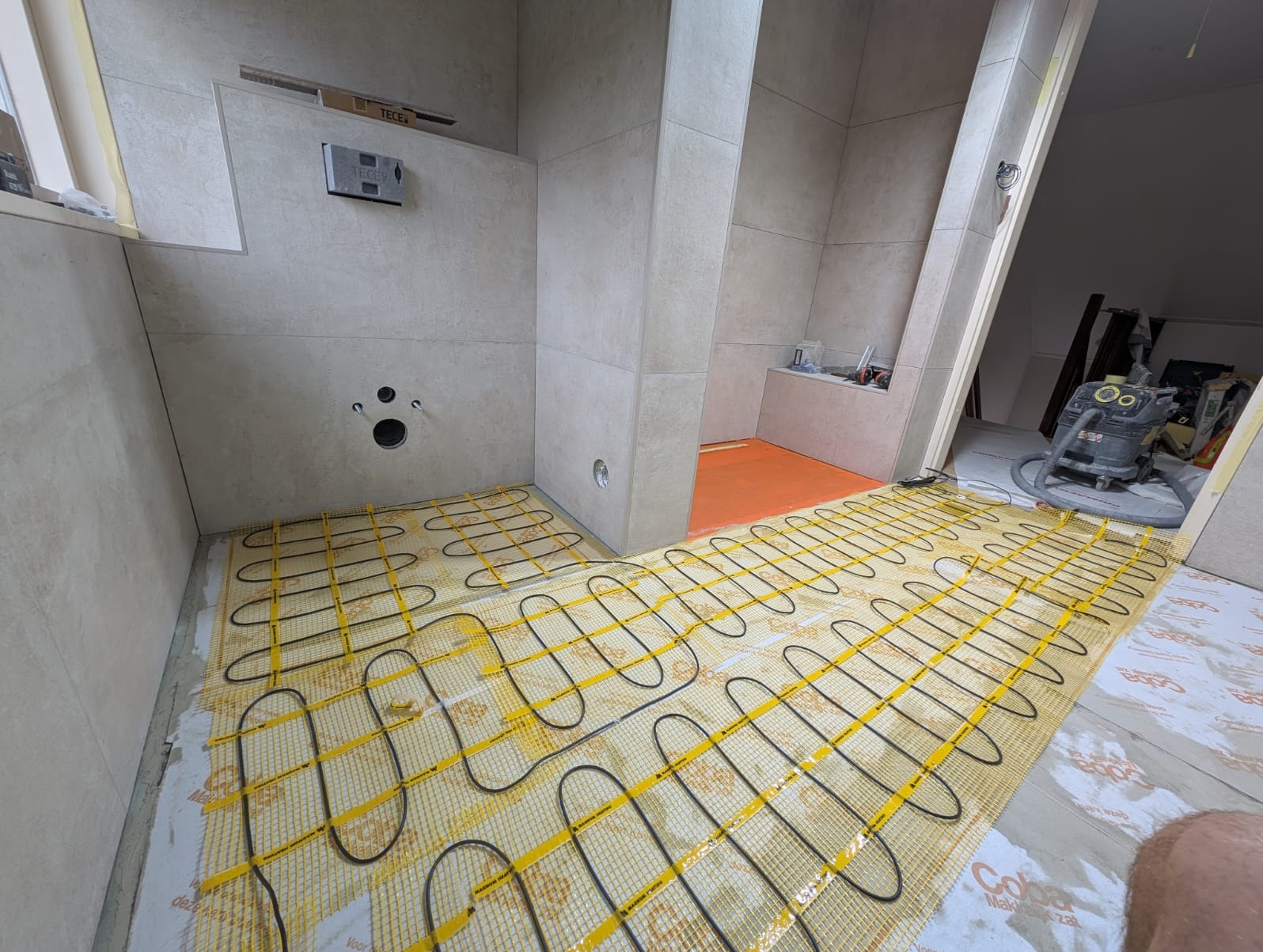 Łazienka w trakcie remontu: ułożona mata grzewcza na podłodze, płytki na ścianach, widoczne przygotowania do dalszych prac wykończeniowych.