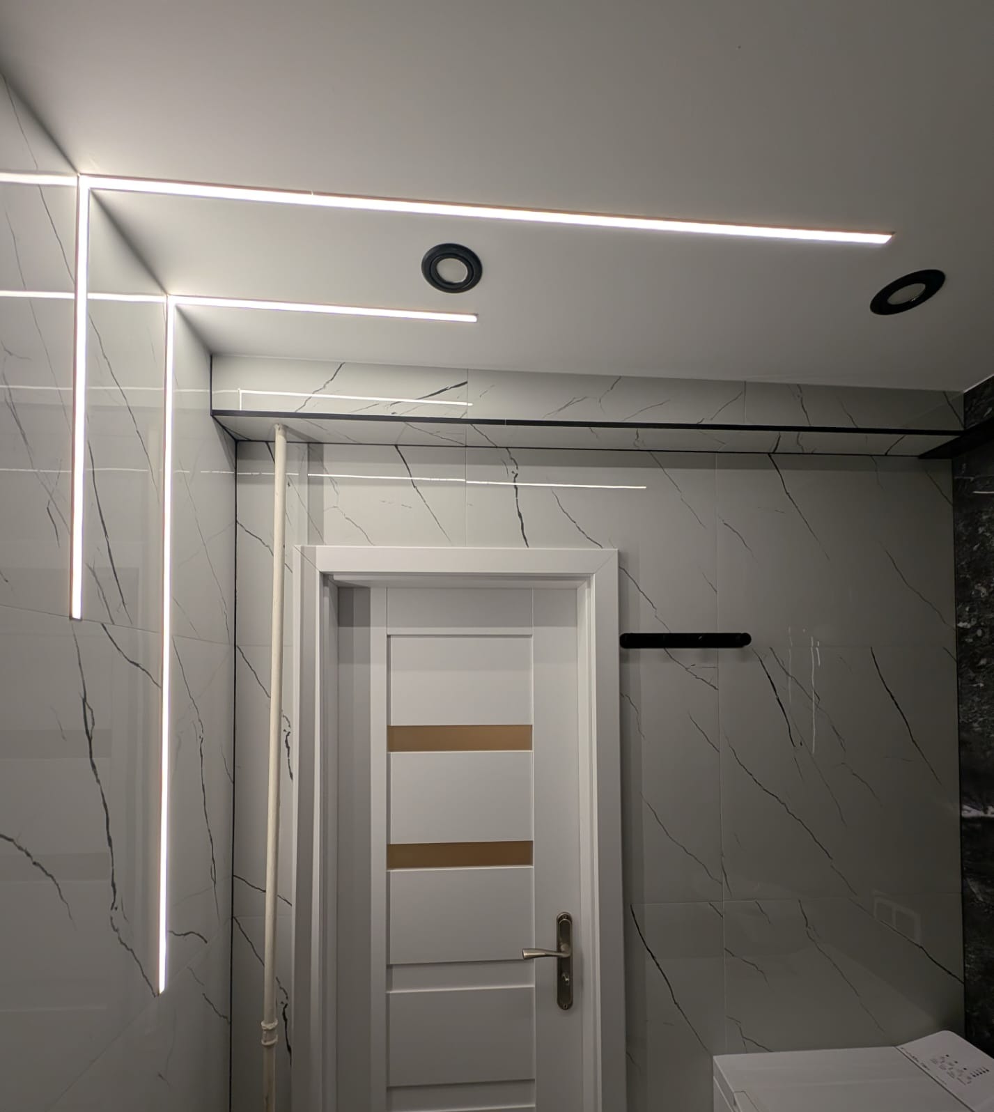 Wykończona łazienka z białymi płytkami imitującymi marmur, oświetleniem LED w suficie i drzwiami z poziomymi paskami. Widoczne czarne halogeny i reling na ręczniki.