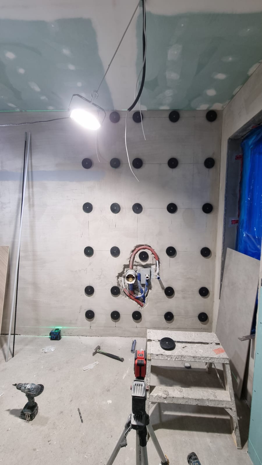 Ściana z zamocowanymi kołkami, widoczne instalacje wodne i elektryczne, narzędzia pracy, poziomica laserowa. Przygotowanie do prac wykończeniowych w pomieszczeniu.