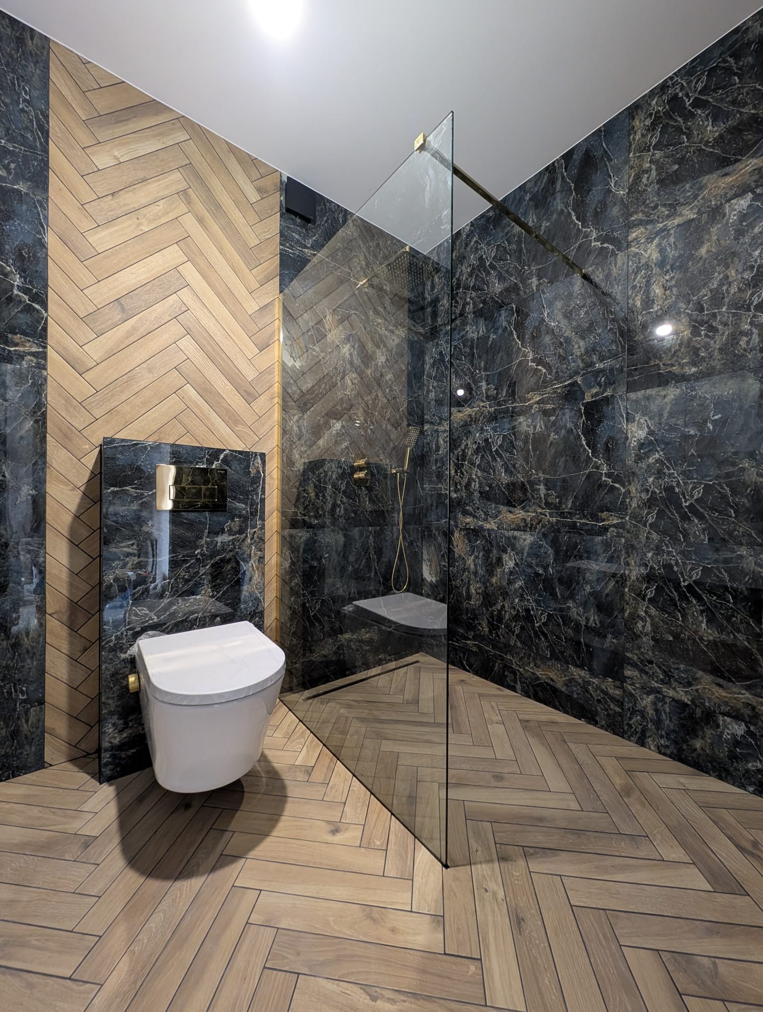 Elegancka łazienka z kabiną prysznicową typu walk-in, wykończona płytkami imitującymi marmur i drewnem ułożonym w jodełkę. Widoczna toaleta podwieszana i złota armatura.