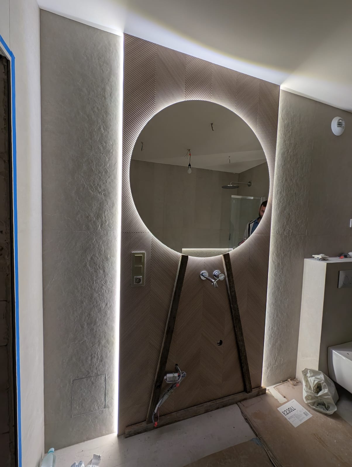 Nowoczesna łazienka w trakcie wykańczania: okrągłe lustro z podświetleniem LED, wzorzysta ściana, widoczne elementy instalacji wodnej i elektrycznej, surowy stan podłogi.