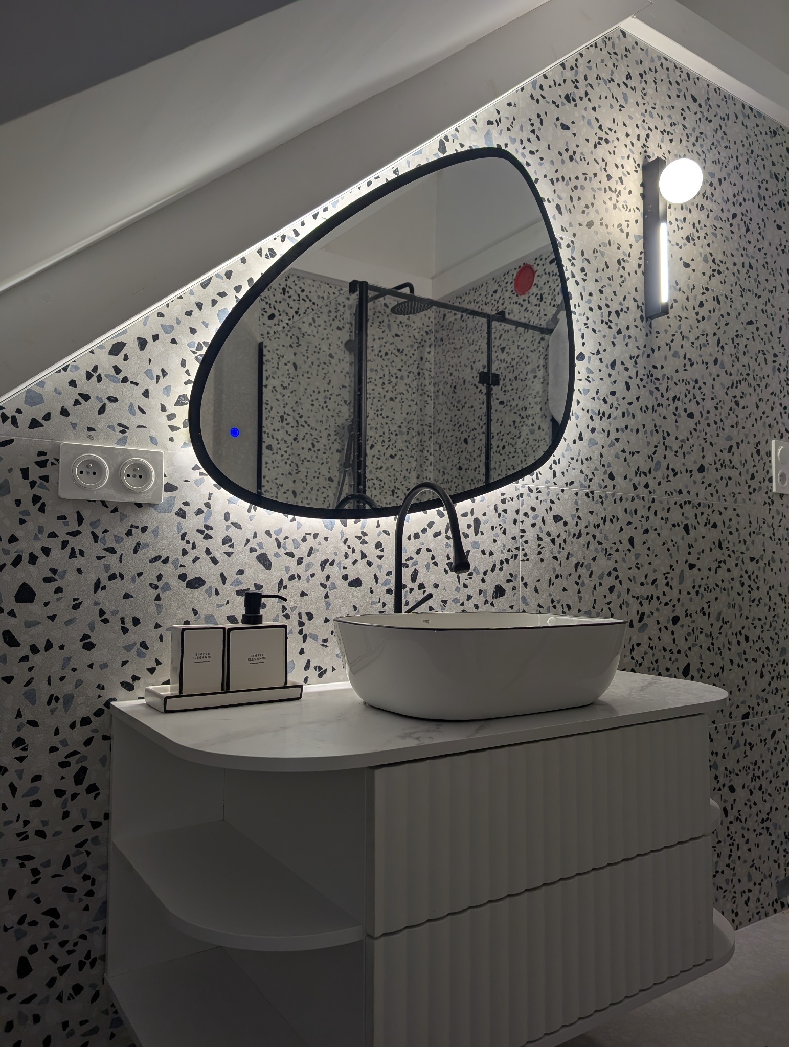 Nowoczesna łazienka z umywalką nablatową, podświetlanym lustrem o nieregularnym kształcie i wzorzystymi płytkami w stylu lastryko na ścianach.