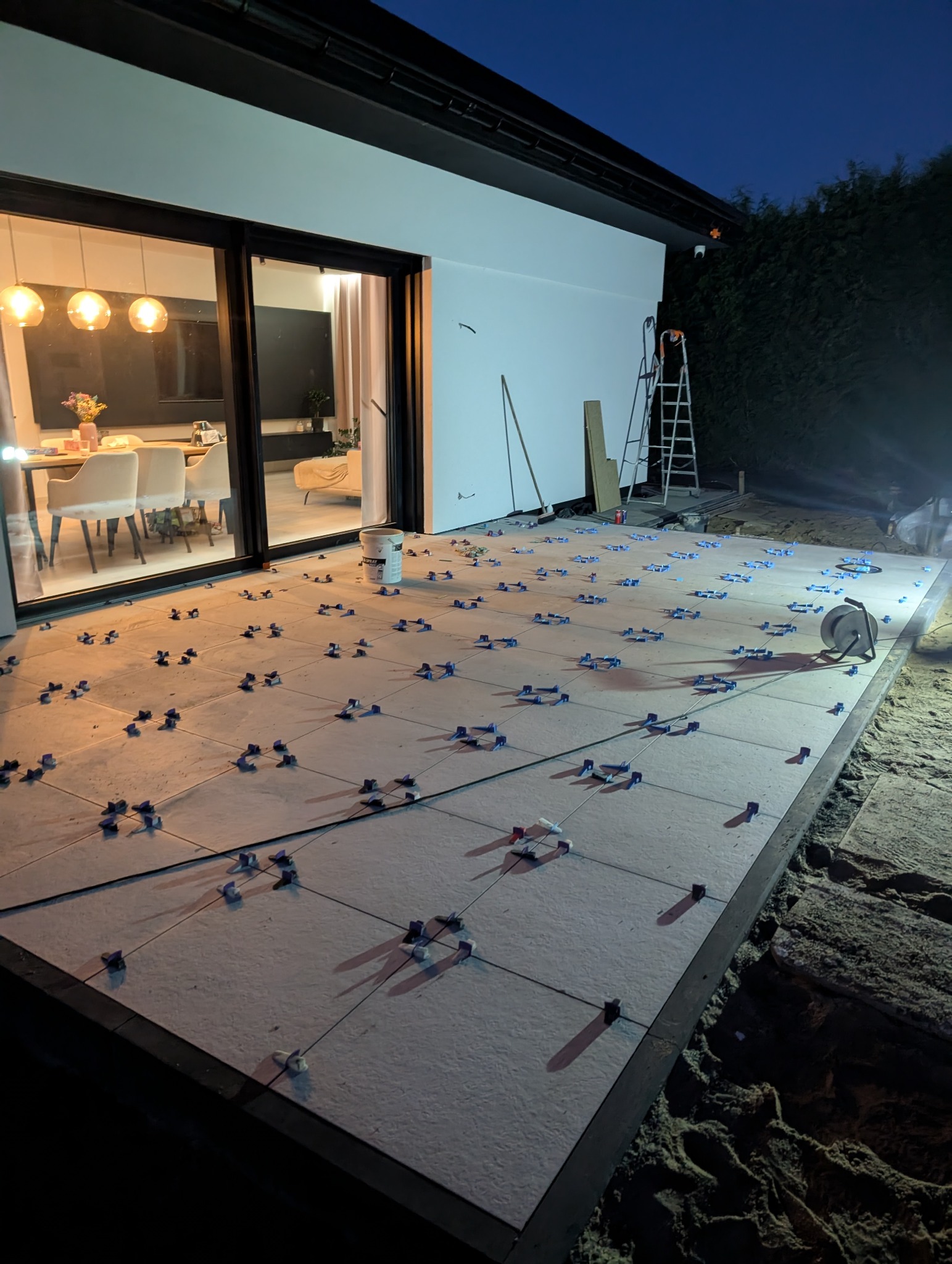 Układanie jasnych płytek na tarasie domu jednorodzinnego z użyciem systemu poziomowania; widoczne narzędzia i drabina. Praca w toku, oświetlenie nocne.