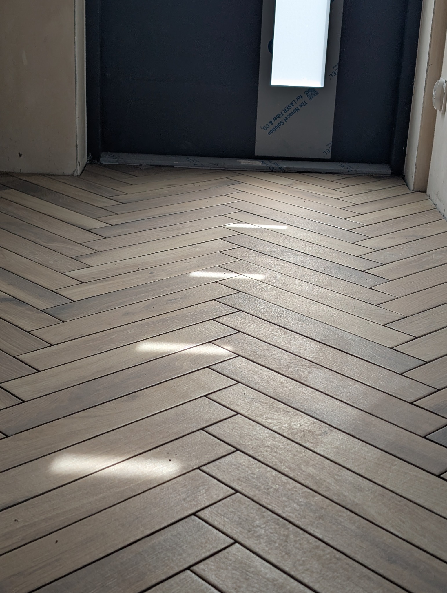 Ułożona w jodełkę podłoga z płytek imitujących drewno, z widocznymi promieniami słońca. W tle fragment ściany i drzwi.