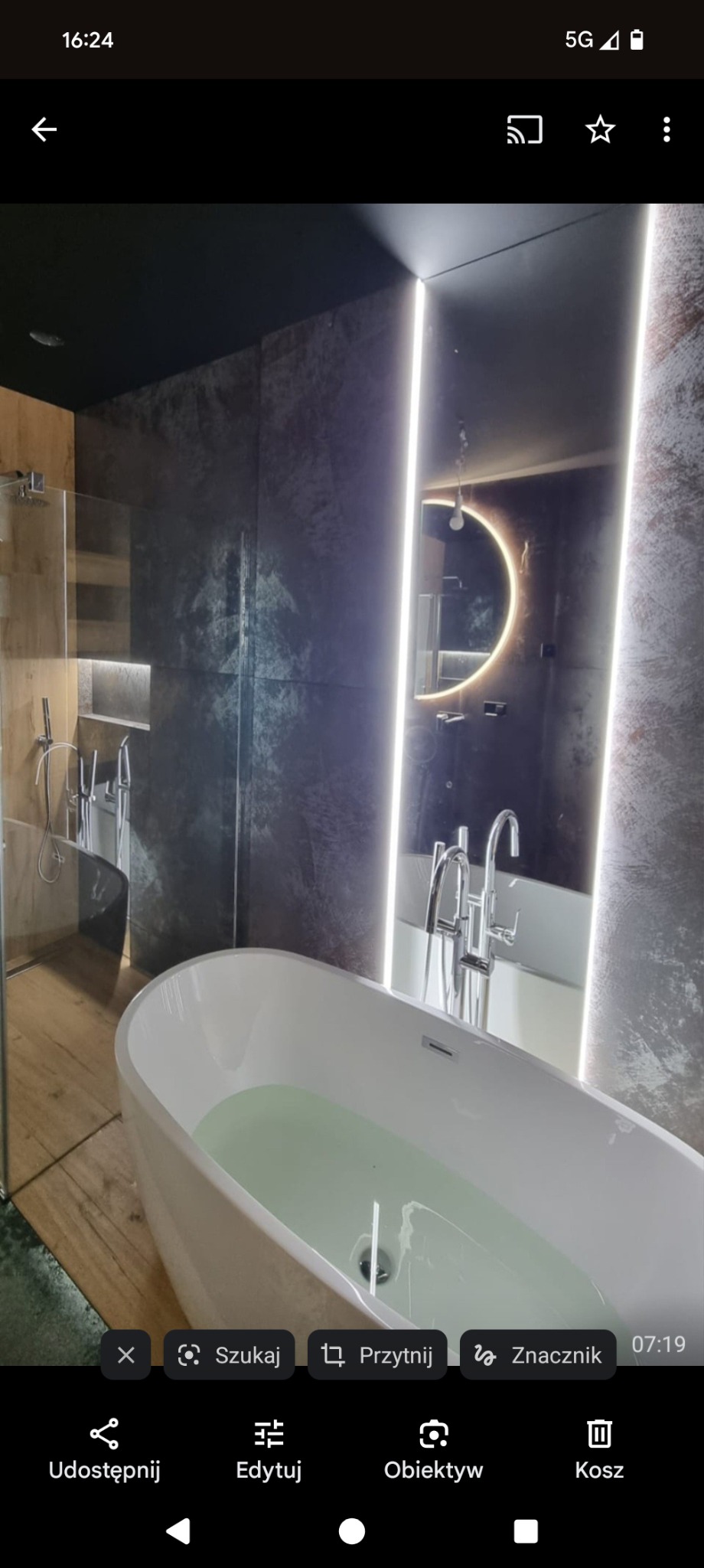 Nowoczesna łazienka z wolnostojącą wanną wypełnioną wodą, podświetlanym lustrem w kształcie półksiężyca i szklaną kabiną prysznicową. Ciemne płytki i drewniane akcenty.