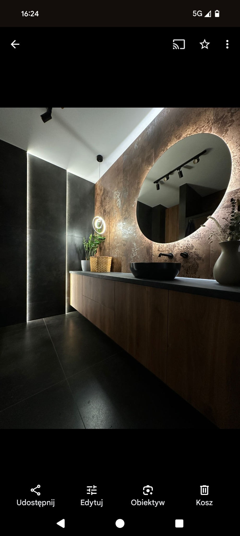 Nowoczesna łazienka z podświetlanym lustrem owalnym, drewnianą szafką pod umywalką i ciemnymi płytkami na ścianach oraz podłodze. Minimalistyczny design z roślinami.