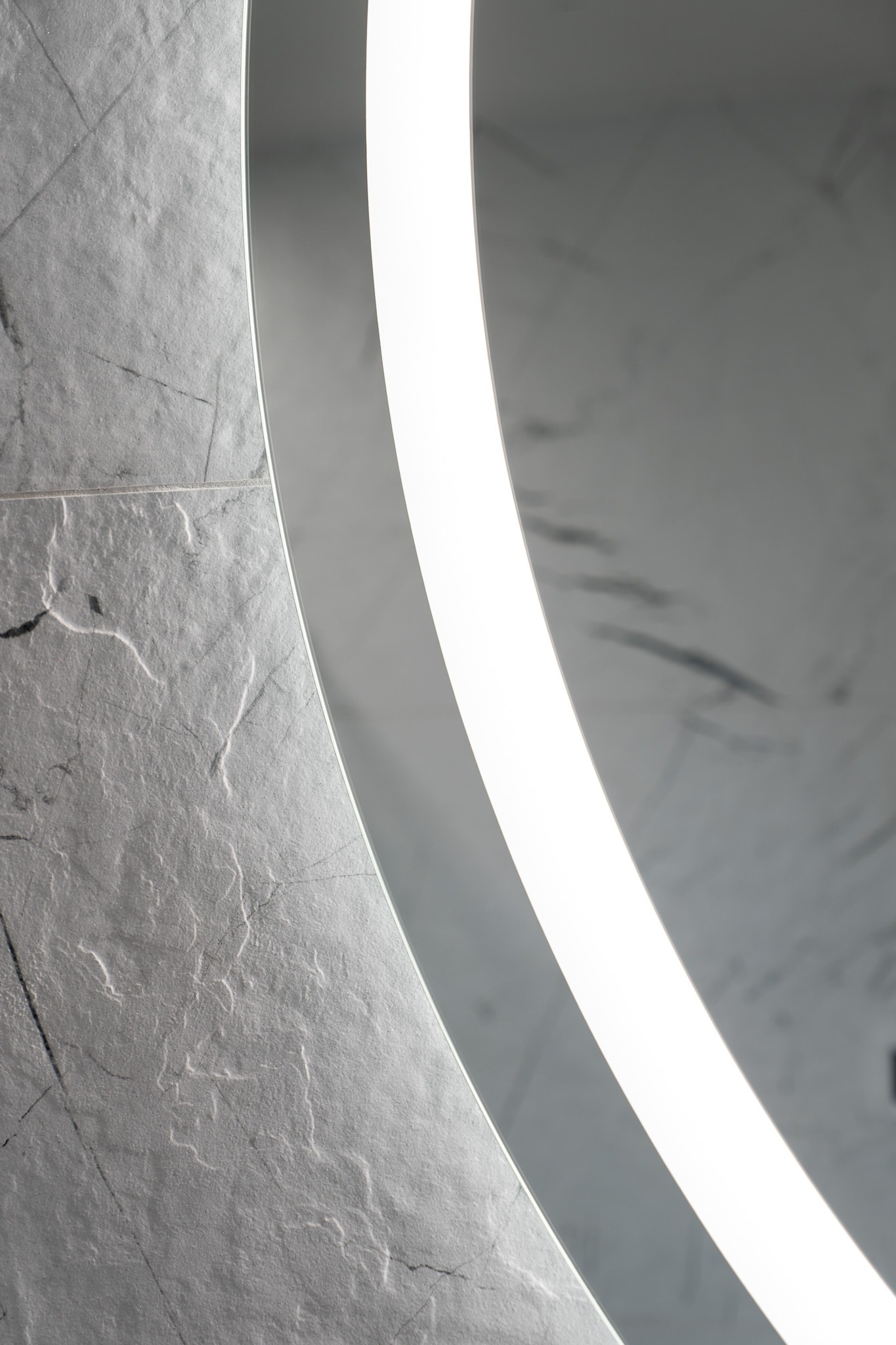 Fragment ściany wyłożonej szarą, kamienną płytką, obok fragment okrągłego lustra z podświetleniem LED.