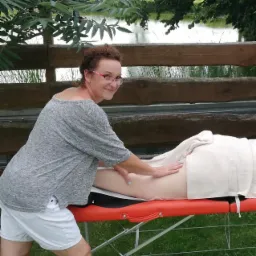 Kobieta w okularach wykonuje masaż uda pacjentki na pomarańczowym stole do masażu na zewnątrz, w tle drewniany płot i staw.