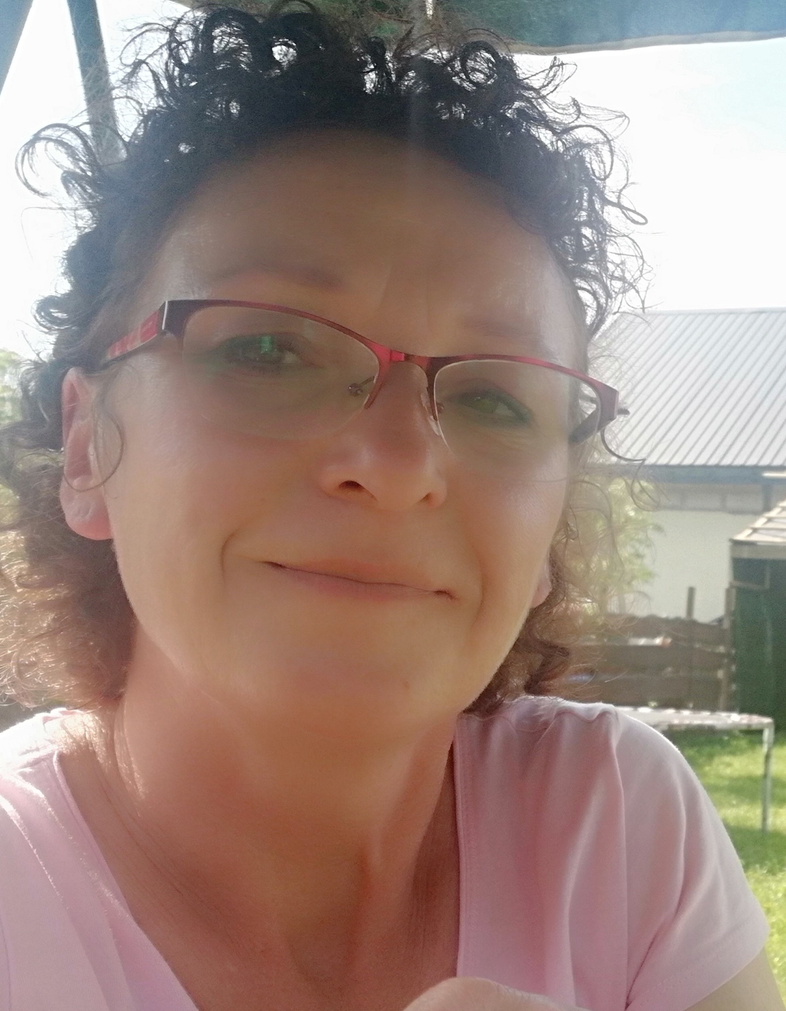 Portret uśmiechniętej kobiety w okularach z kręconymi włosami na tle ogrodu z budynkiem gospodarczym