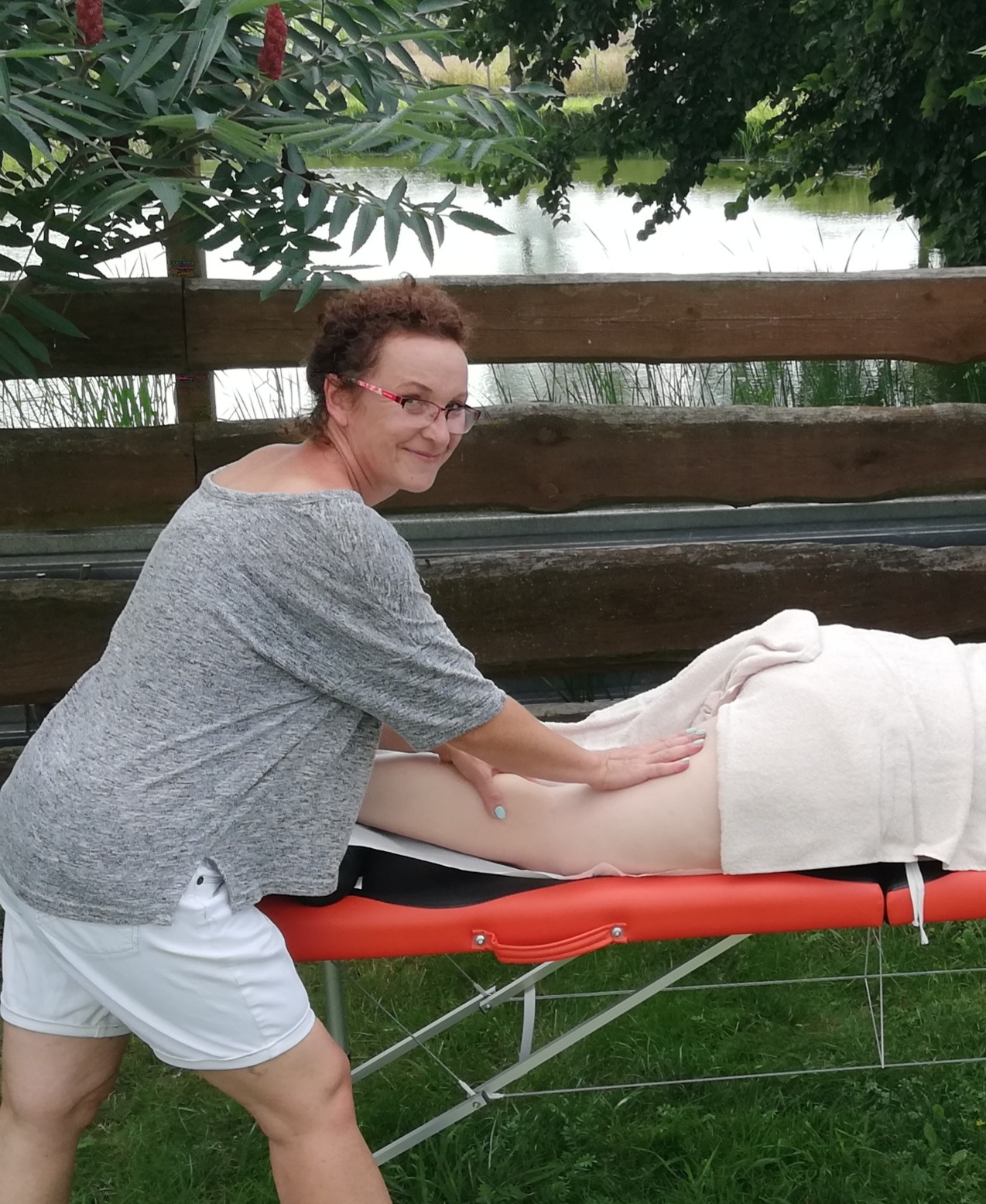 Kobieta w okularach wykonuje masaż uda pacjentki na pomarańczowym stole do masażu na zewnątrz, w tle drewniany płot i staw.