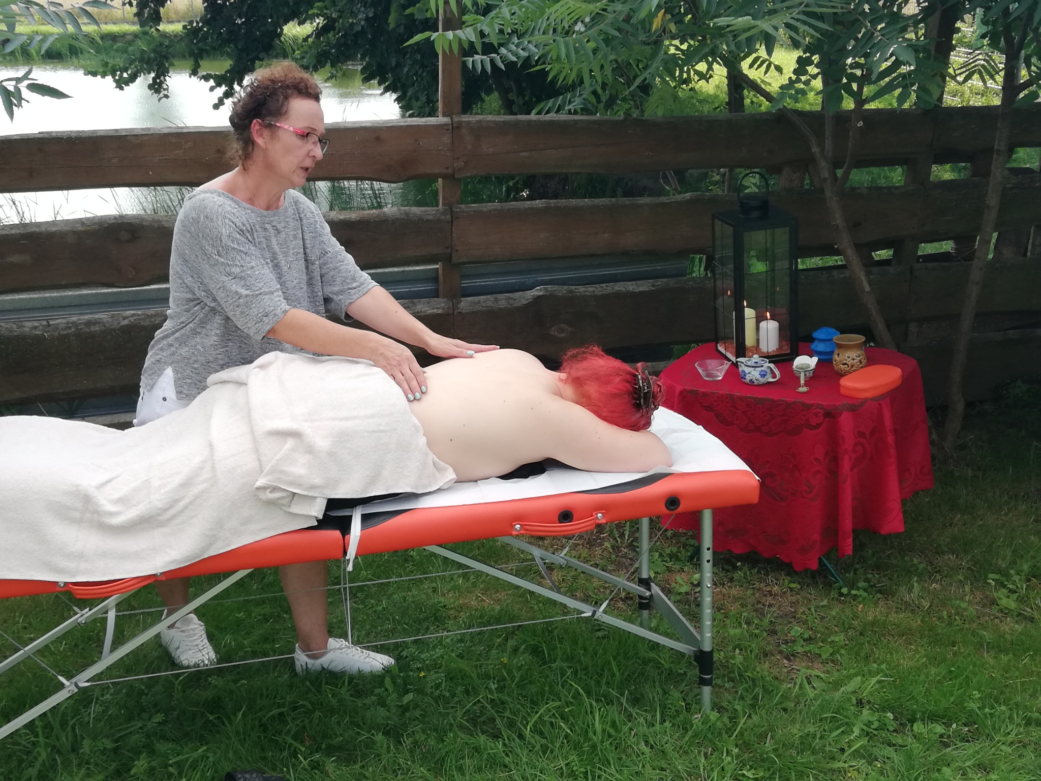 Kobieta wykonuje masaż pleców na pomarańczowym stole do masażu, w tle drewniany płot, jezioro i stolik z lampionem i świecami.