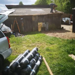 Mężczyzna na dachu drewnianej szopy, prawdopodobnie przygotowuje podłoże pod nowe pokrycie dachowe. Na trawie leżą rolki papy dachowej, obok zaparkowany samochód z otwartym bagażnikiem.