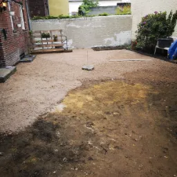 Przygotowanie terenu pod trawnik lub kostkę brukową, widoczne częściowo wyr&oacute;wnane podłoże z piasku i ziemi, paleta z kwiatami przy ścianie budynku.