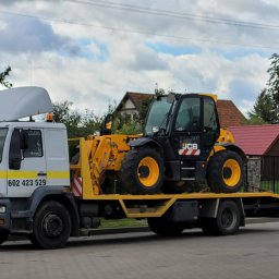 TRANSPORTER Piotr Danowski - Żółto-czarny podnośnik teleskopowy JCB przewożony na białej lawecie MAN z napisem 'Pomoc Drogowa' na tle zabudowań jednorodzinnych.