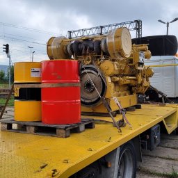 TRANSPORTER Piotr Danowski - Żółta platforma ciężarówki z załadowanym agregatem prądotwórczym Caterpillar i dwoma paletami z beczkami w kolorach czerwonym i żółtym na tle torów kolejowych i pochmurnego nieba.