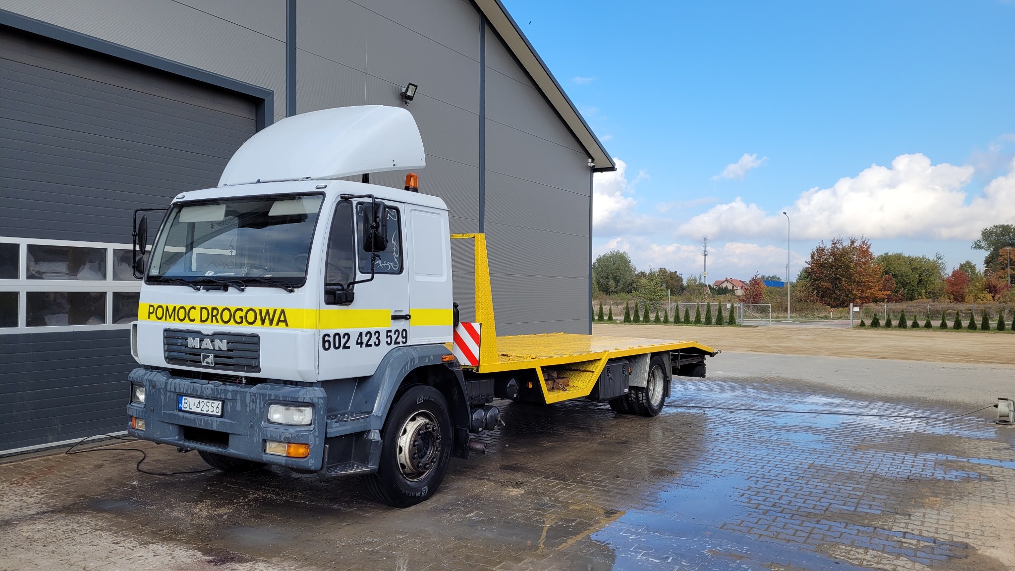Żółta laweta MAN z napisem 'Pomoc Drogowa' zaparkowana przed szarą halą przemysłową w słoneczny dzień.