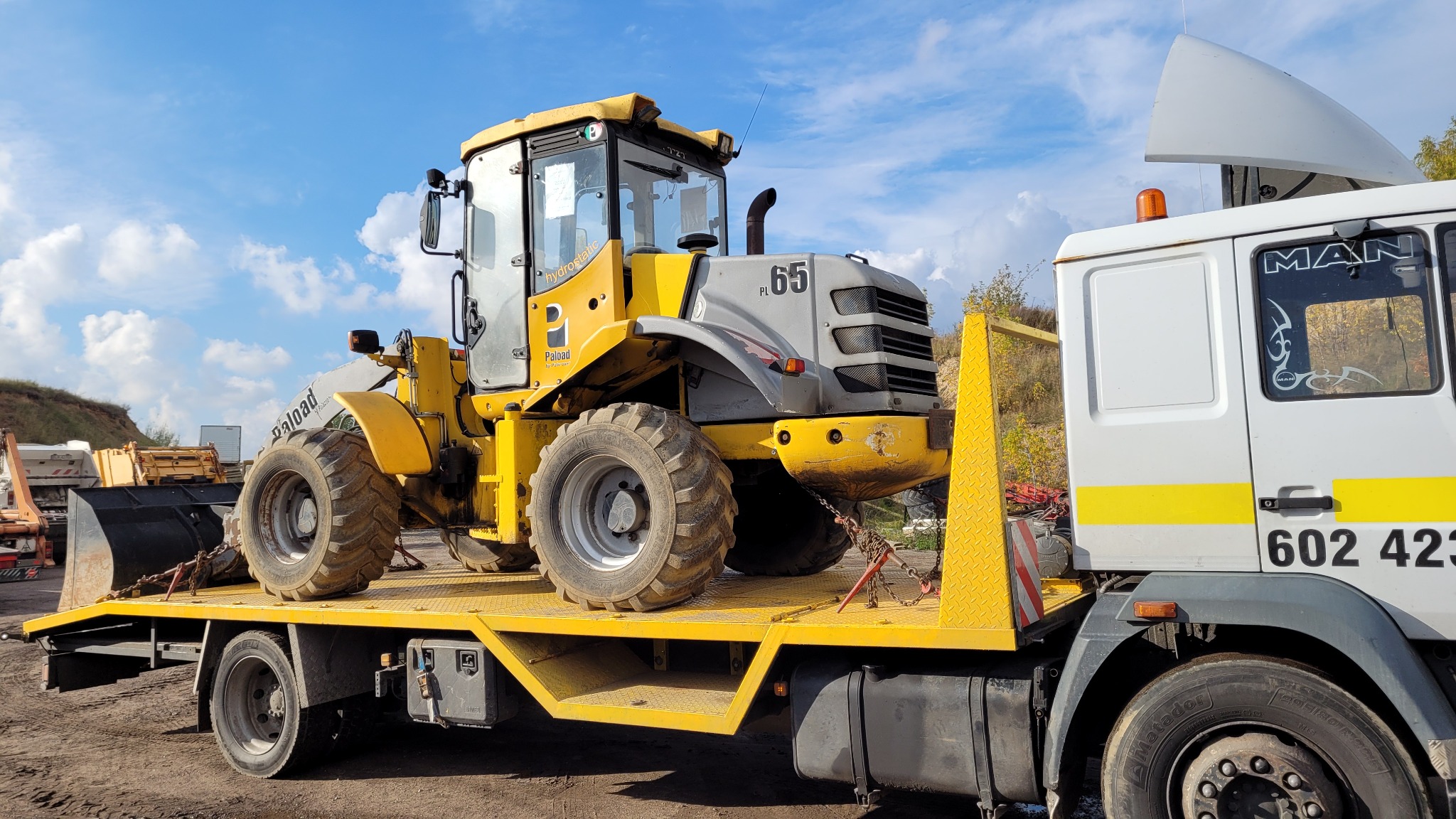 Żółto-szara ładowarka Paload 65 PL transportowana na żółtej lawecie MAN, zabezpieczona łańcuchami z czerwonymi napinaczami, na tle błękitnego nieba z chmurami.