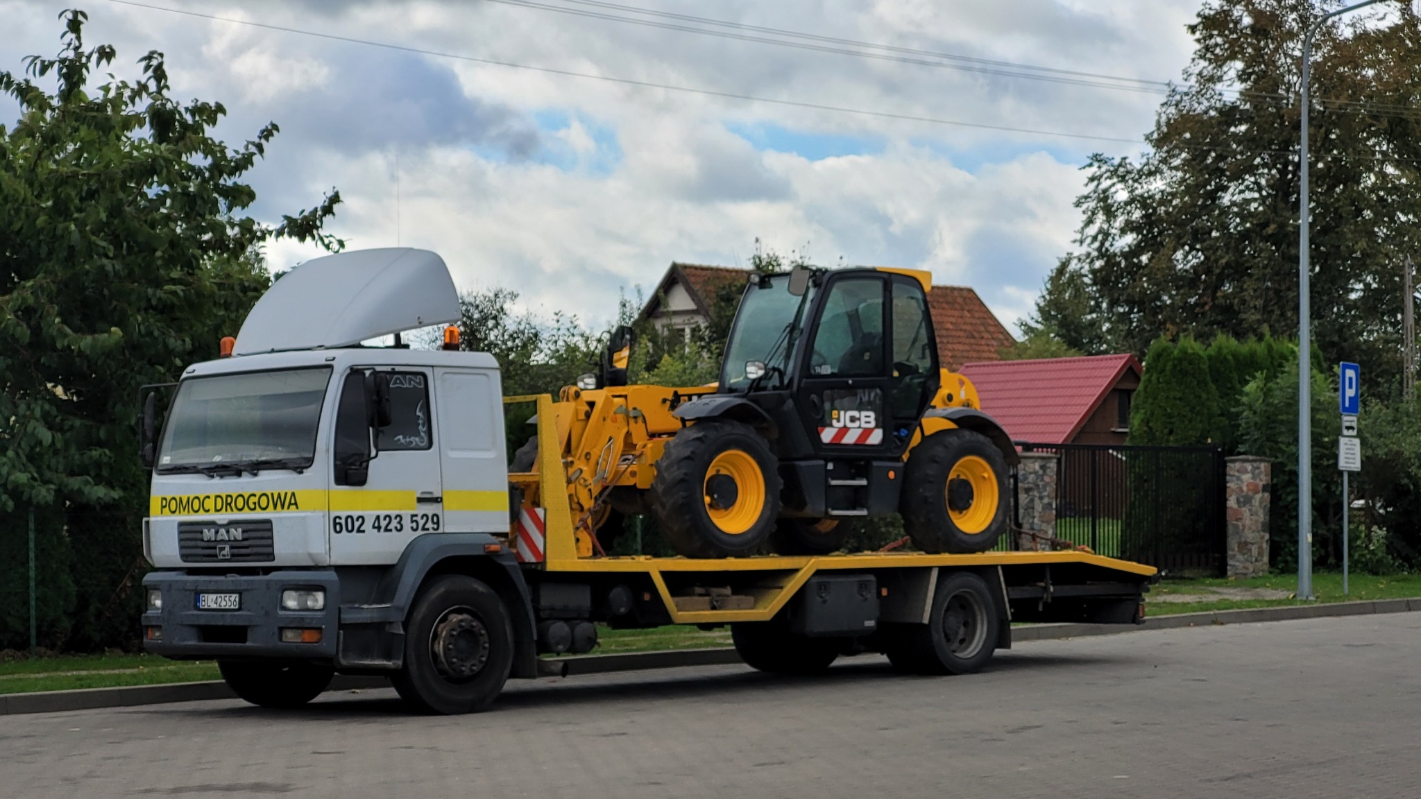 Żółto-czarny podnośnik teleskopowy JCB przewożony na białej lawecie MAN z napisem 'Pomoc Drogowa' na tle zabudowań jednorodzinnych.