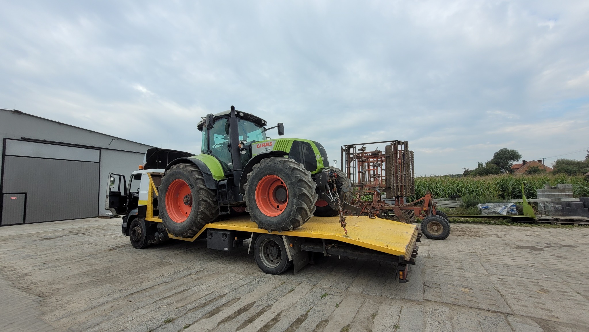 Zielony traktor marki Claas Axion 850 na żółtej platformie lawety, z narzędziem rolniczym w tle, na betonowym placu.