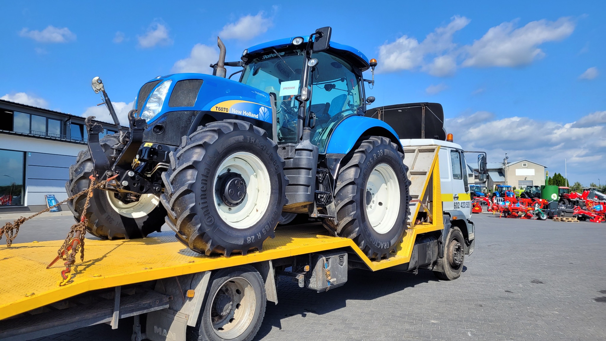 Niebieski traktor New Holland T6070 zabezpieczony łańcuchami na żółtej platformie lawety MAN, widok z boku, w tle plac z maszynami rolniczymi i budynek z dużymi oknami, błękitne niebo z chmurami.