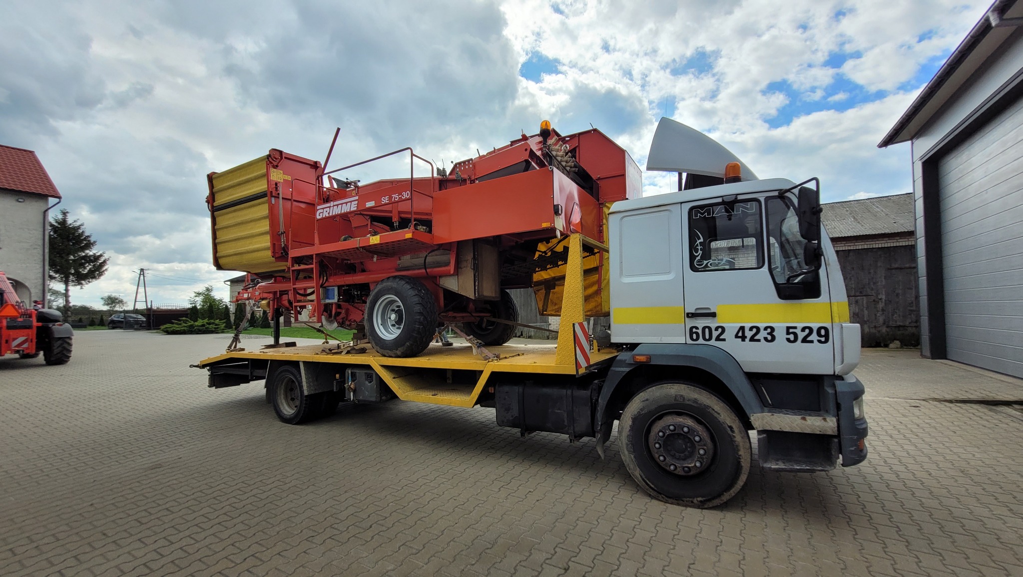 Ciężarówka MAN z żółtą platformą przewozi kombajn ziemniaczany Grimme SE 75-30 na tle budynków gospodarczych i pochmurnego nieba.