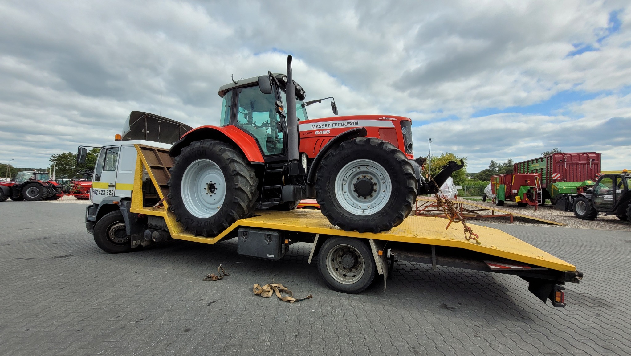 Czerwony traktor Massey Ferguson Dyno 6465 zabezpieczony łańcuchami na żółtej platformie lawety z białym ciągnikiem, widok z boku na szarym brukowanym placu z innymi maszynami rolniczymi w tle.