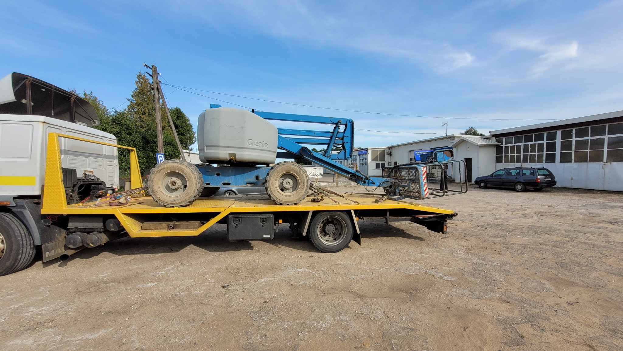 Żółta laweta z niebieskim podnośnikiem Genie na platformie, zaparkowana na placu przed budynkiem z rzędem okien.