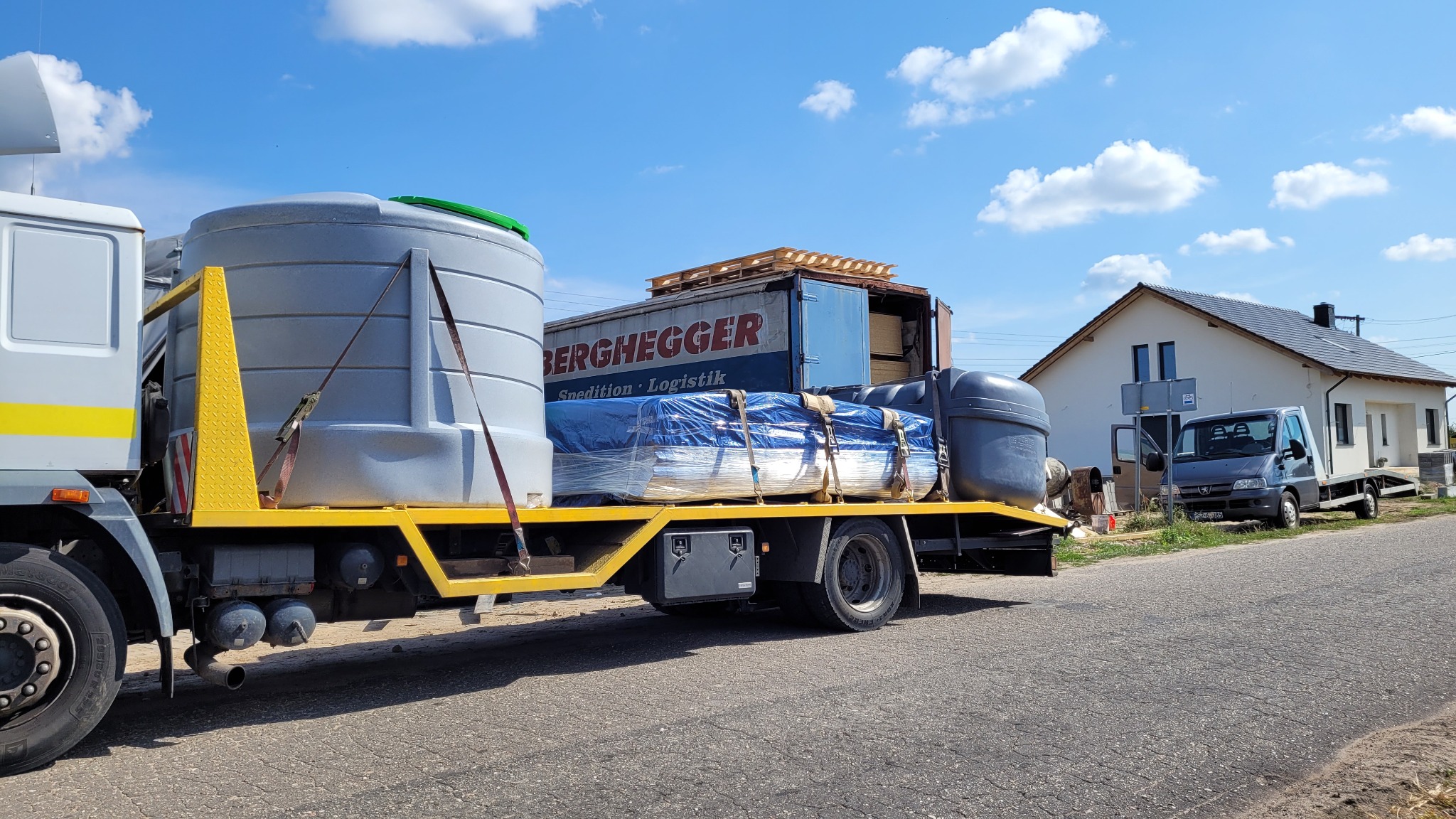 Ciężarówka z żółtą platformą transportuje dwa szare zbiorniki z zielonymi pokrywami, przykryte folią materiały budowlane i palety, zaparkowana na poboczu drogi w słoneczny dzień, w tle dom...