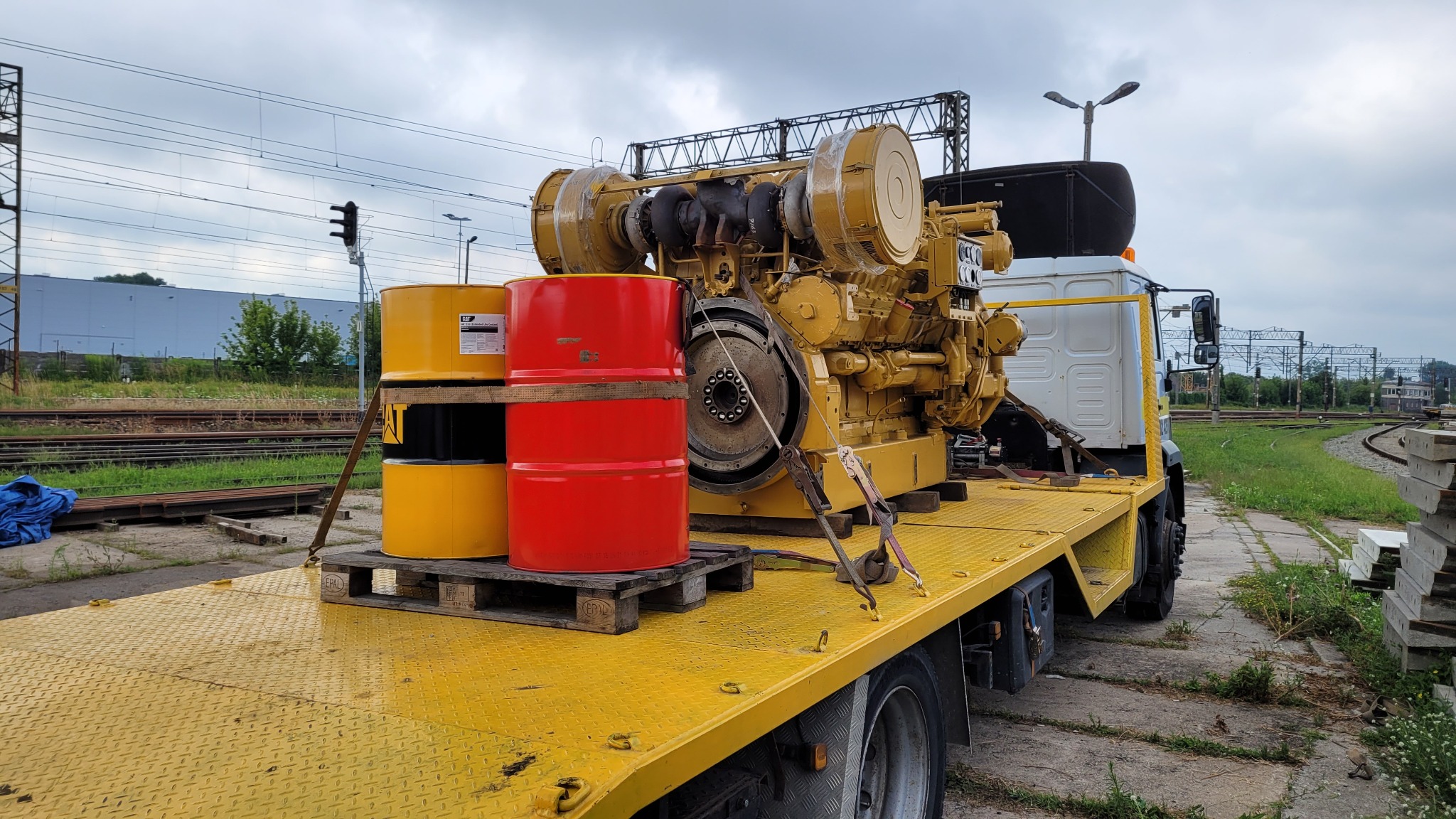 Żółta platforma ciężarówki z załadowanym agregatem prądotwórczym Caterpillar i dwoma paletami z beczkami w kolorach czerwonym i żółtym na tle torów kolejowych i pochmurnego nieba.