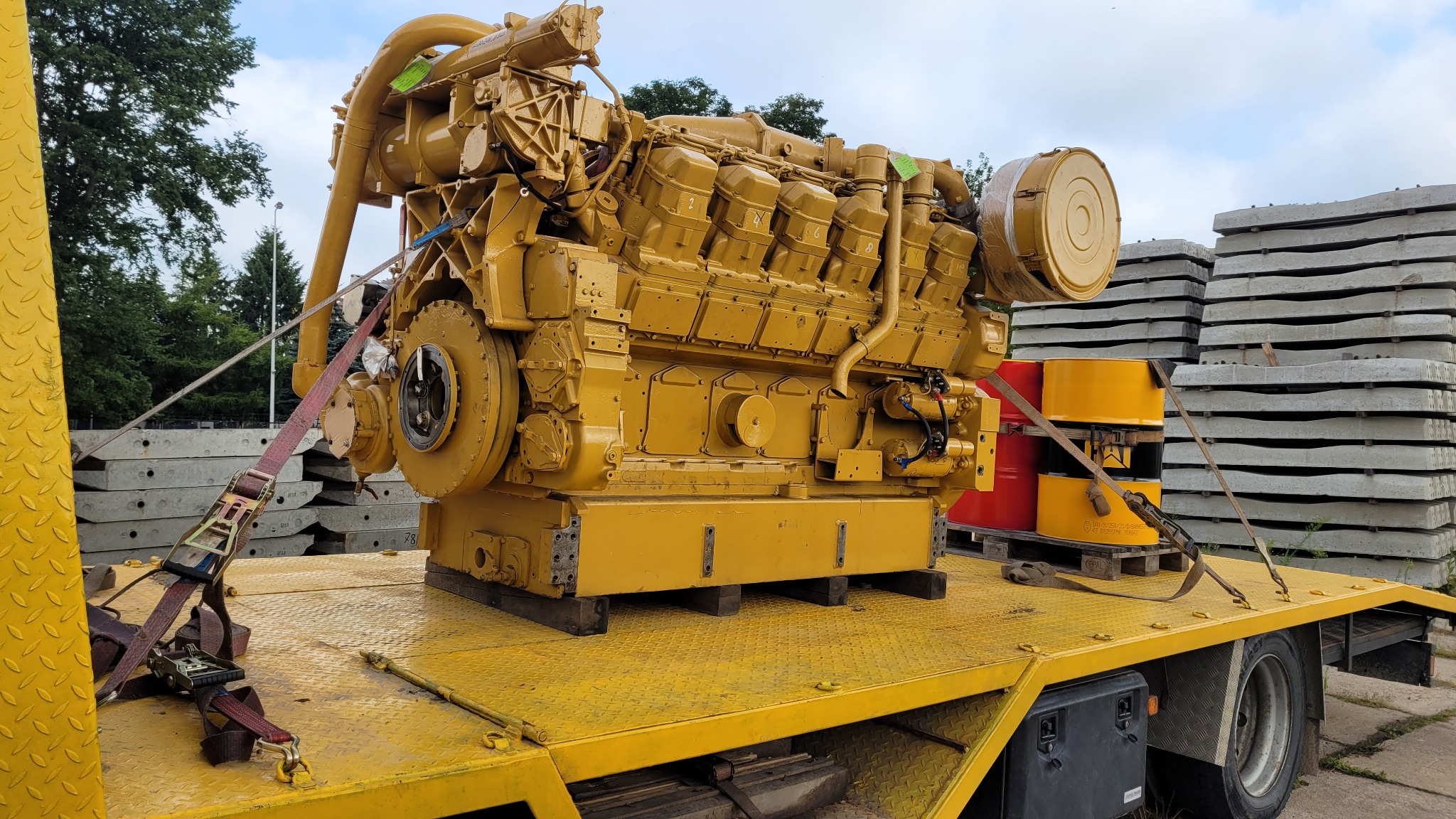 Żółty, przemysłowy silnik spalinowy zabezpieczony pasami transportowymi na platformie lawety, w tle betonowe płyty i beczki.