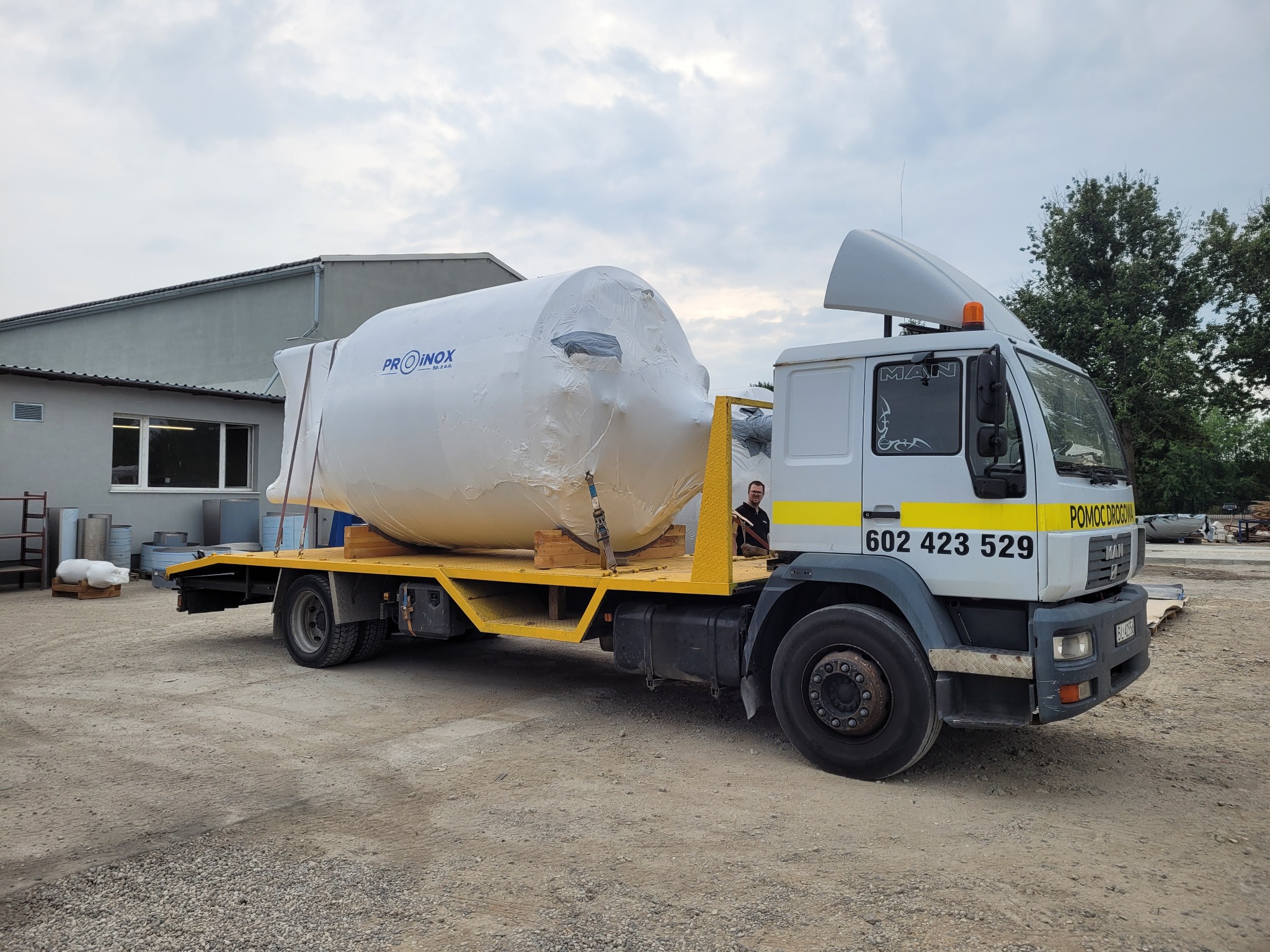Laweta z załadowanym, owiniętym folią zbiornikiem ze stali nierdzewnej z logo PROINOX, na tle budynku przemysłowego i gruntu żwirowego, widoczny kierowca w kabinie.