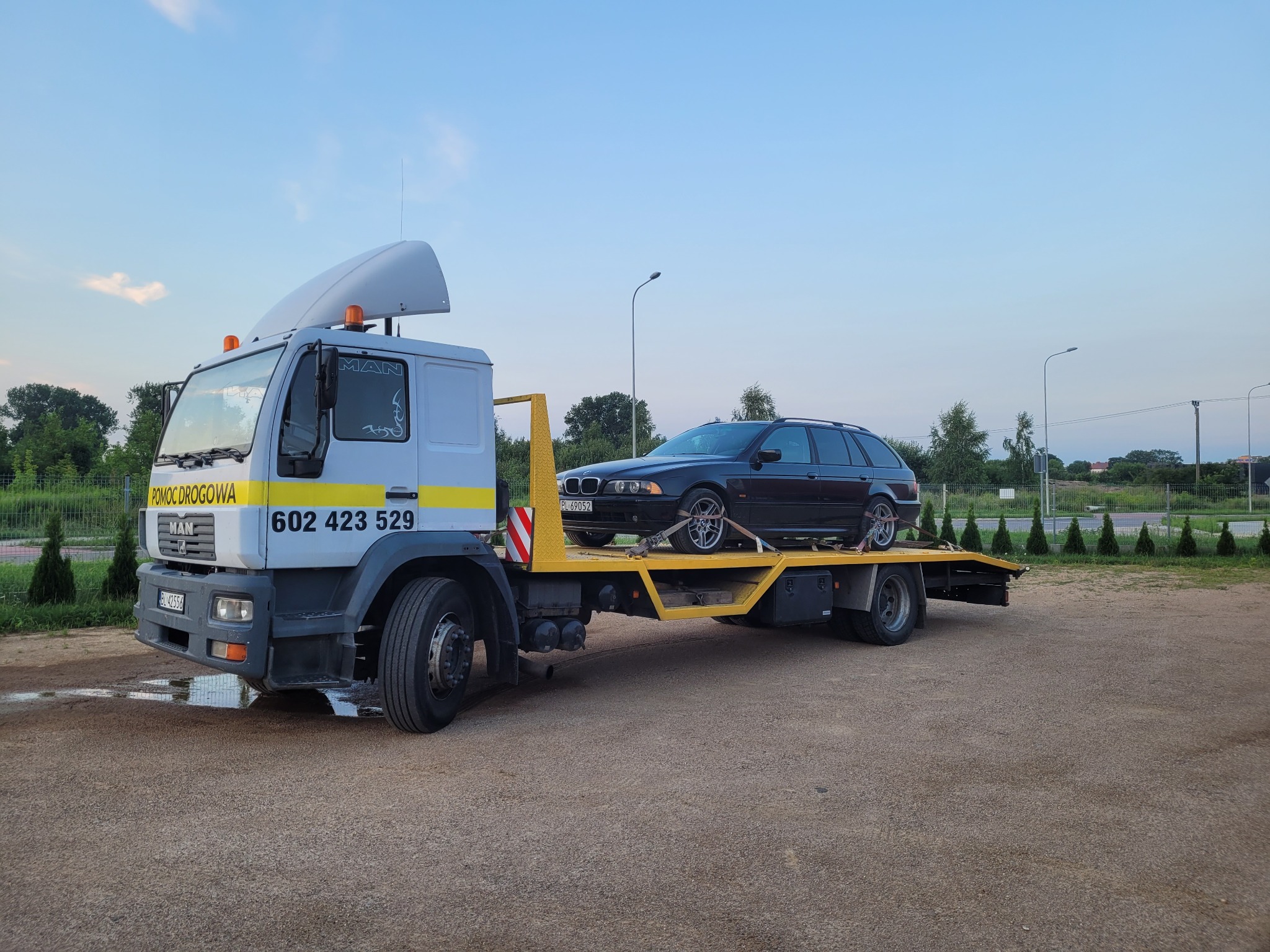 Czarna limuzyna BMW kombi zabezpieczona pasami na żółtej platformie lawety MAN z napisem POMOC DROGOWA i numerem telefonu na kabinie, na szarym placu z zielenią w tle.