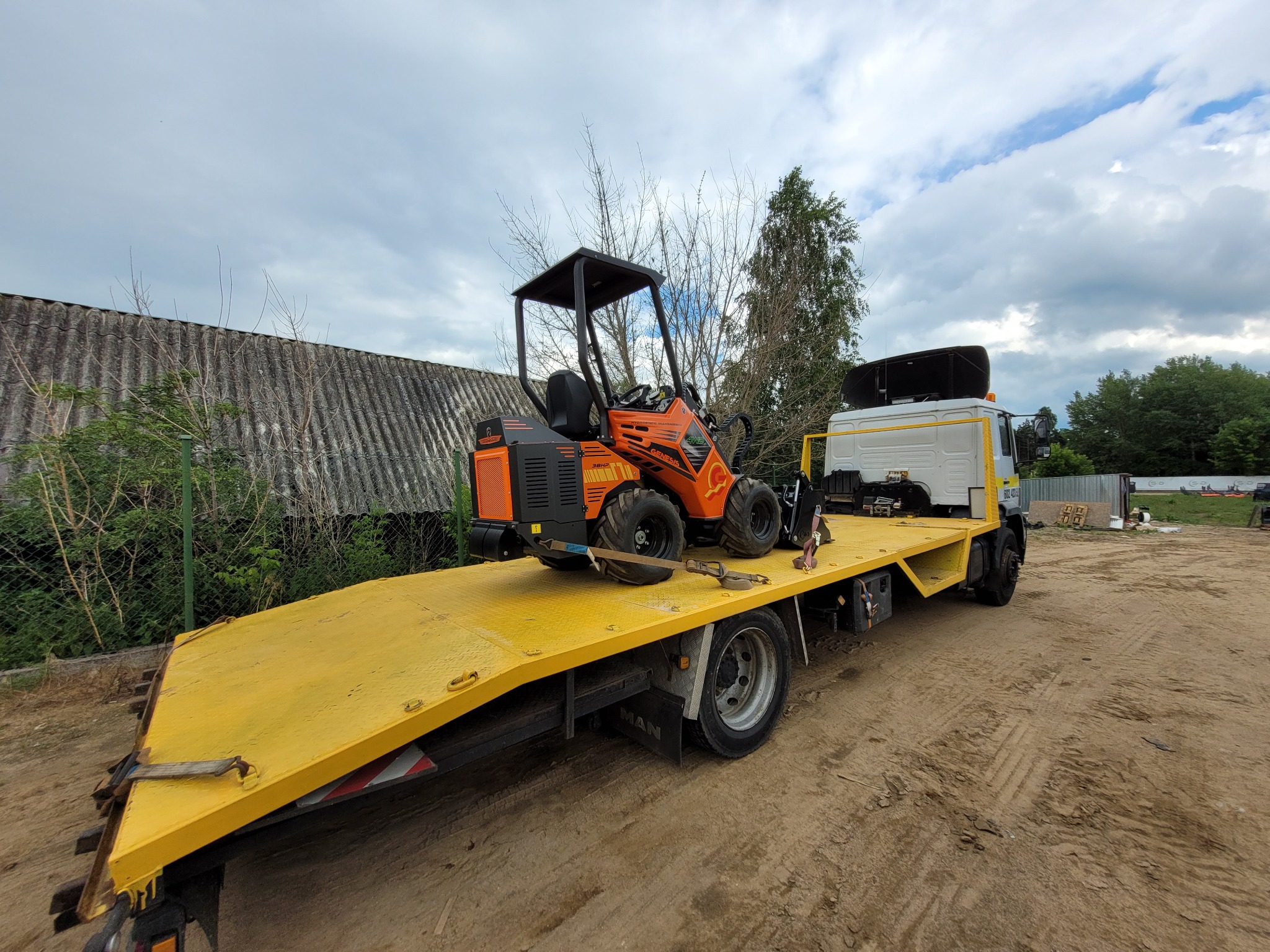 Pomarańczowa miniładowarka Genesis 36H2 zabezpieczona pasami na żółtej platformie lawety MAN, widok z boku, na tle szarego dachu i pochmurnego nieba.