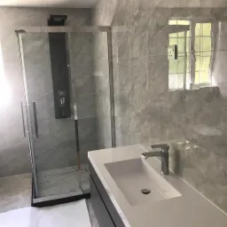 Wykończona łazienka w Puławach z szarą marmurkową glazurą, kabiną prysznicową z hydromasażem i umywalką wpuszczaną w blat z szafką, widok na okno z zielenią na zewnątrz.