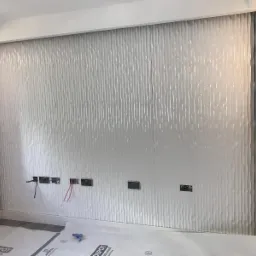 Ściana z dekoracyjną, białą, falistą okładziną, z przygotowanymi otworami na gniazdka elektryczne, zabezpieczona folią ochronną na podłodze.