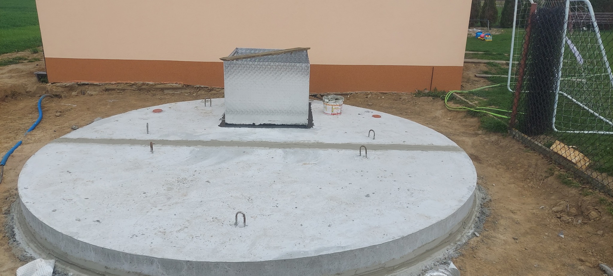 Betonowa konstrukcja szamba z metalową pokrywą na posesji, widoczne elementy montażowe i ślady prac budowlanych, tło stanowi budynek i ogrodzenie.