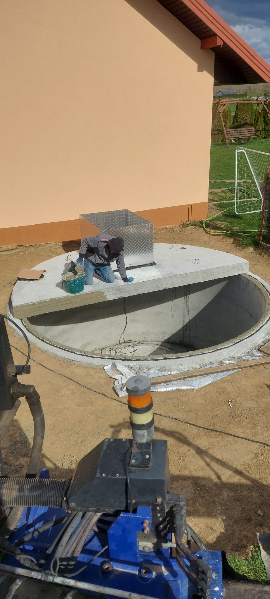 Pracownik uszczelnia betonowy zbiornik szamba przy budynku. Widoczny sprzęt budowlany i narzędzia. Praca na zewnątrz w słoneczny dzień.