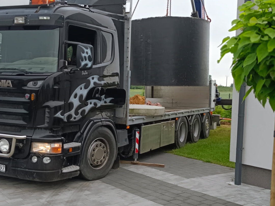 Czarna ciężarówka z dźwigiem transportuje betonowy krąg studzienny na palecie. Widoczny fragment posesji i zieleń. Transport kręgów studziennych.