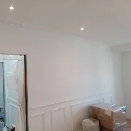 Białe wnętrze w trakcie wykańczania, widoczne listwy dekoracyjne na ścianach i suficie, kartony zabezpieczone folią, oraz otwarte przejście do pomieszczenia z widocznym bałaganem i narzędziami...