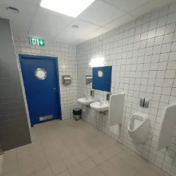 Wnętrze publicznej toalety: białe kwadratowe płytki na ścianach, jasna podłoga z płytek, niebieskie drzwi z okrągłym okienkiem, umywalki, pisuary i dozowniki mydła.