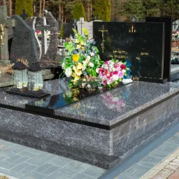 Granitowy pomnik z czarną tablicą inskrypcyjną i bukietami kwiatów na cmentarzu, widoczne lampiony.