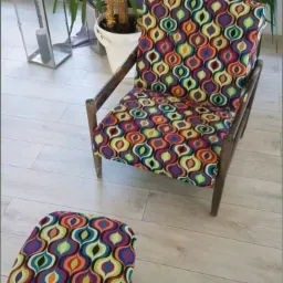 Tapicerowane krzesło i podnóżek w stylu retro, z wzorzystą tkaniną w kolorowe łezki, na tle jasnych paneli podłogowych i rośliny doniczkowej.