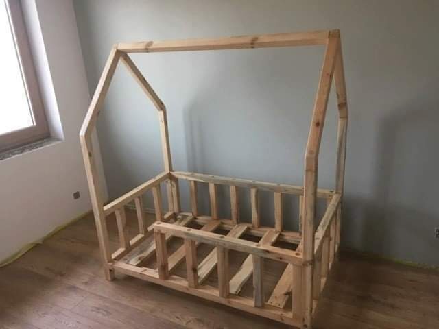Surowa rama drewniana w kształcie domku, prawdopodobnie łóżka dziecięcego, z widocznymi niedoskonałościami obróbki drewna i surowym wykończeniem, ustawiona na drewnianej podłodze w pokoju z szarą...