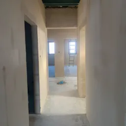 Korytarz w trakcie tynkowania maszynowego gipsowego; widoczne gładkie ściany, surowy strop i drabina w głębi pomieszczenia.
