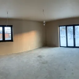 Wnętrze w stanie deweloperskim po nałożeniu gładzi gipsowej, widoczne ślady po pracy na podłodze, okno z widokiem na drewniany dom i zasypany śniegiem ogród, oraz szklane drzwi balkonowe. W rogu...