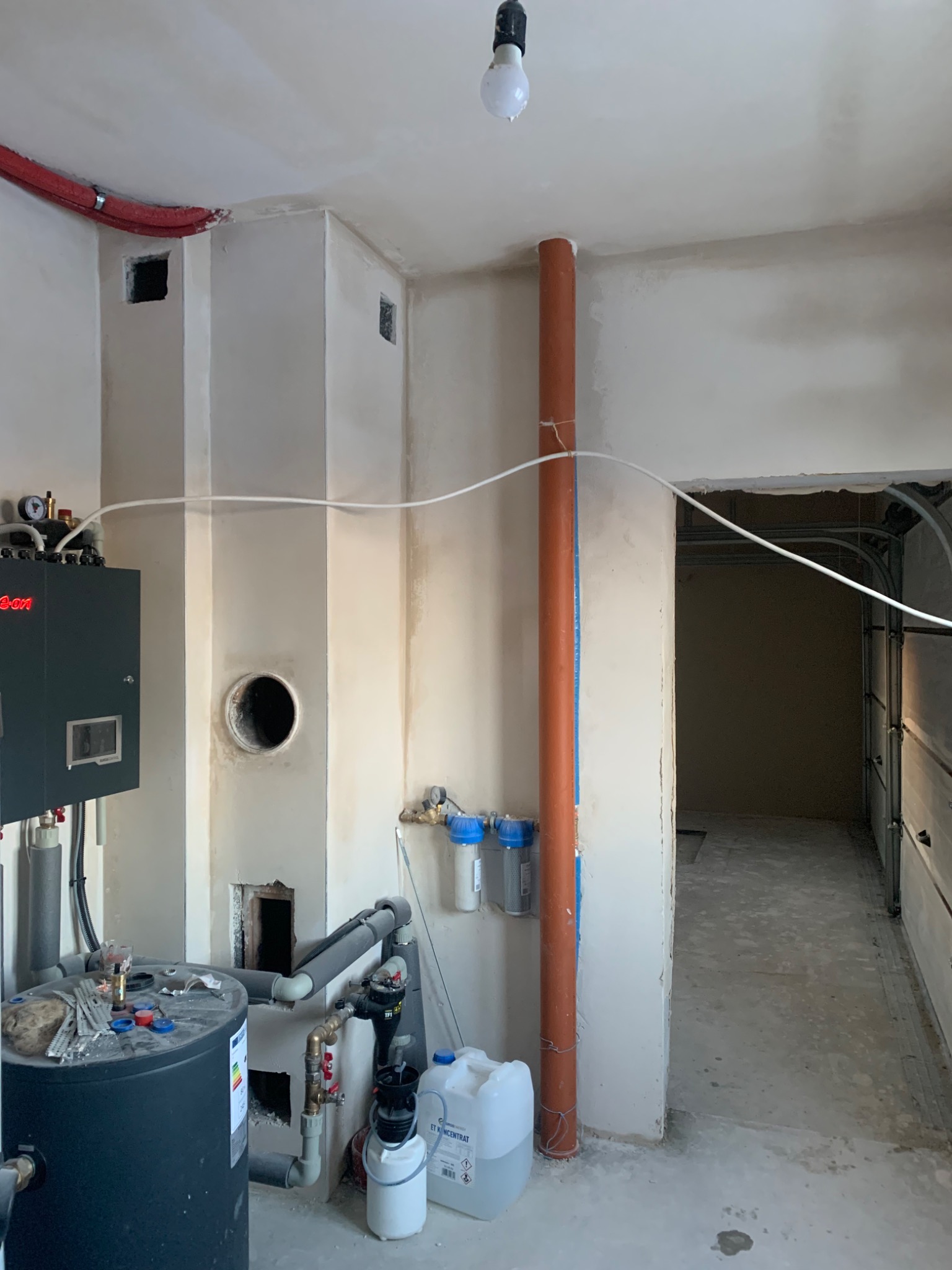 Surowy stan wnętrza po tynkowaniu maszynowym gipsowym, widoczne instalacje hydrauliczne i otwór wentylacyjny w ścianie, sufit z surowym wykończeniem i wiszącą żarówką, podłoga betonowa przed wylewką.
